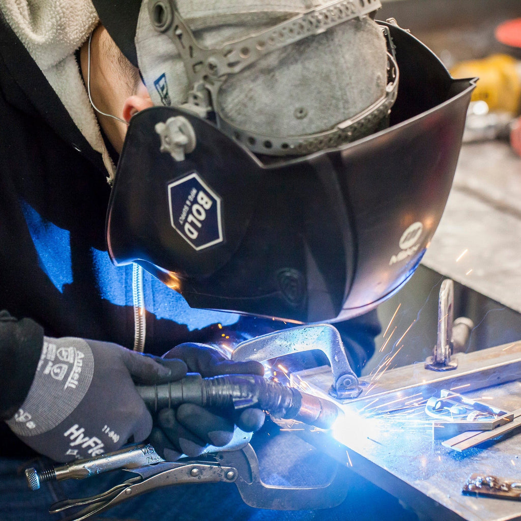 Close up of a person wearing welding gear and welding a piece of steel. A bright blue light emits from the end of the welding tool.