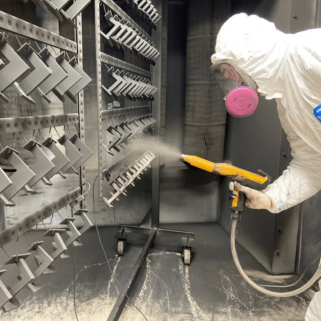 A person in PPE uses a paint sprayer to coat pieces of metal hanging from racks