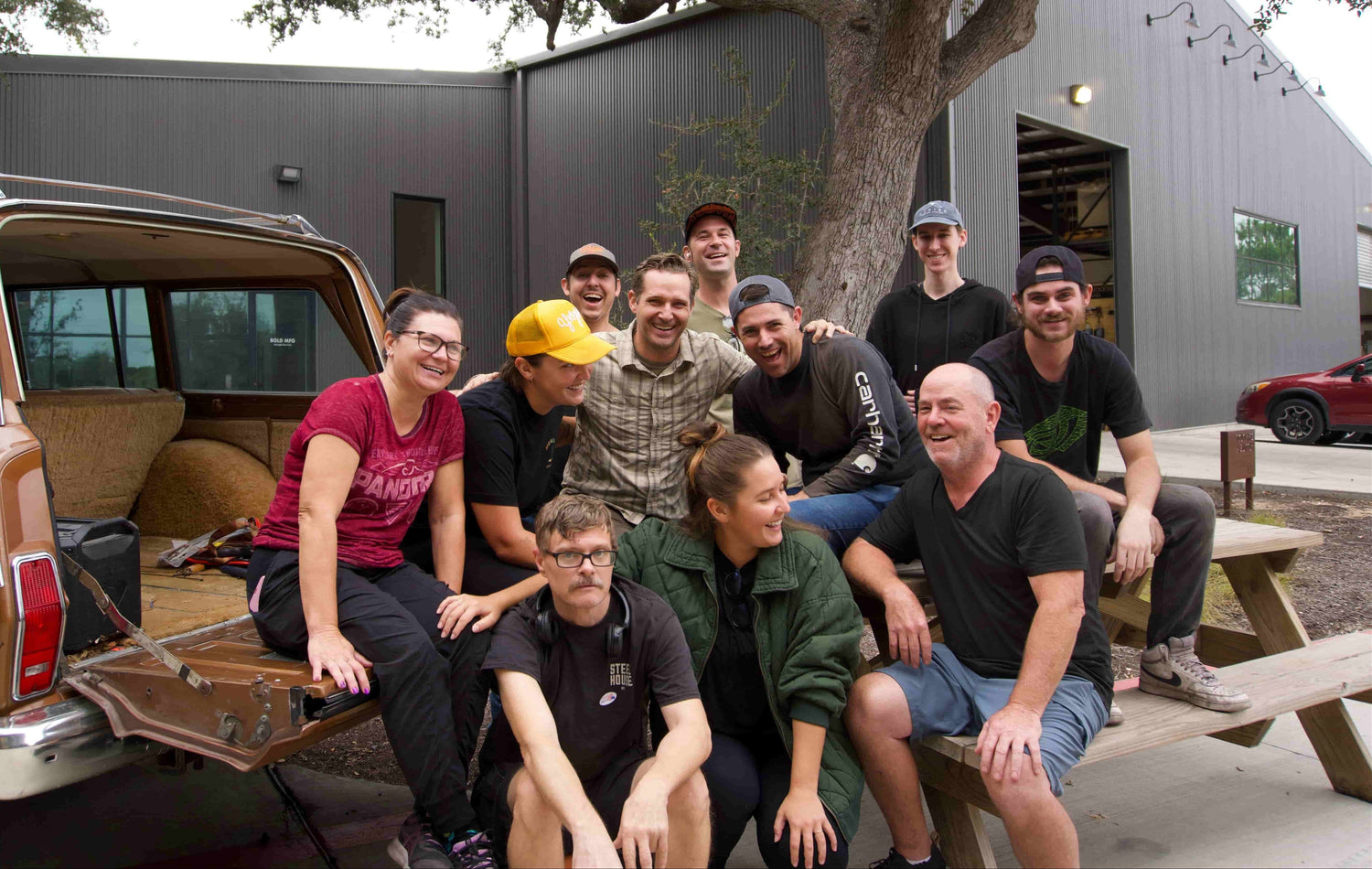 A group of people gathers and smiles on a picnic table and car tailgate. They're outdoors, in front of a grey building and near a parked vehicle.